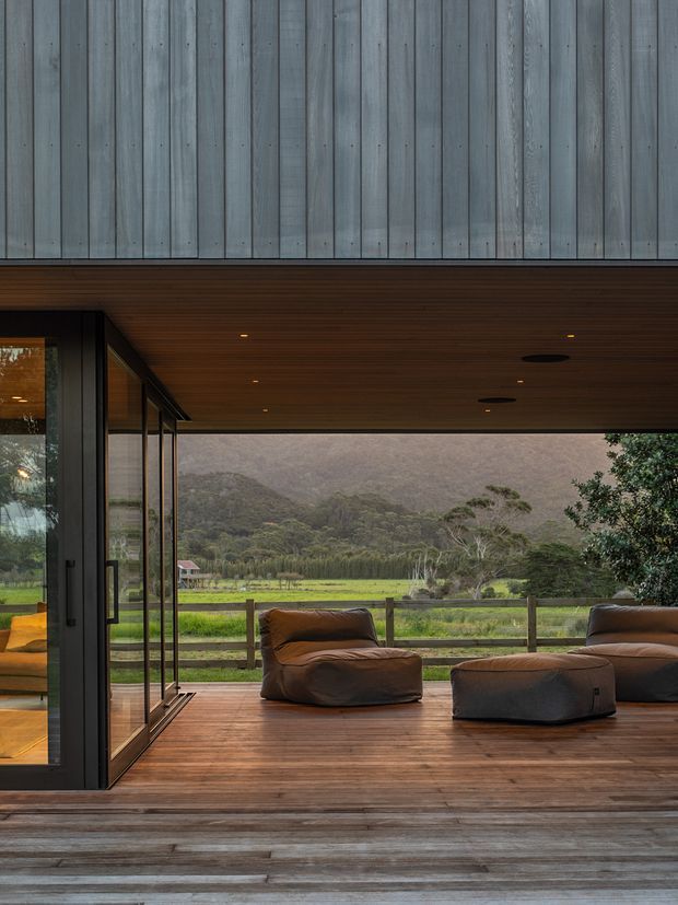 A covered outdoor deck or terrace with warm timber decking and a timber-lined ceiling, furnished with low brown lounge chairs. Large black-framed glazed sliding doors open to the interior, while a rural green landscape and forested mountain backdrop are visible beyond the simple timber railing. The image conveys a refined, warm indoor-outdoor connection.