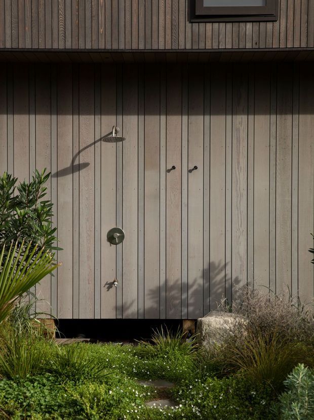 Close exterior view of a timber-clad outdoor shower enclosure, with vertical weathered wood boards, dark trim and a small upper window. A wall-mounted shower fixture and controls are set into the cladding, surrounded by dense ornamental landscaping and stepping stones.