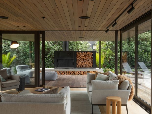 A warm, contemporary living room opening onto a timber deck and lush garden through extensive floor-to-ceiling glazing. The room features a pale upholstered sofa arrangement, dark structural framing, a timber-lined ceiling with recessed and track lighting, and a central black fireplace/media unit with an integrated firewood storage niche.