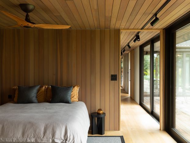 Warm, timber-lined bedroom with a low-profile bed, neutral bedding and dark accent pillows. A ceiling fan and track lighting complement the wood-plank ceiling and walls, while full-height glazed doors open onto a bright adjoining corridor or outdoor-facing space.