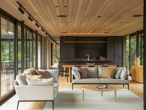 A warm, open-plan living room with a modern kitchen at the rear. The space features extensive floor-to-ceiling glazing along one side, a timber-lined ceiling with recessed and track lighting, neutral upholstered sofas, a light timber coffee table, and dark built-in kitchen cabinetry with a compact island.