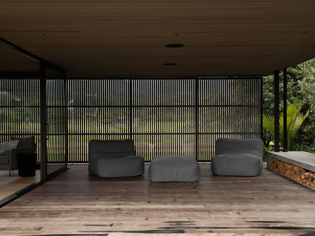 A covered outdoor lounge deck with a timber ceiling and weathered timber flooring. Floor-to-ceiling black-framed glazing and vertical slatted screening enclose the space, with low grey lounge chairs and an ottoman arranged around the open deck. Lush greenery is visible beyond the screening, and a stone-faced built-in edge runs along the right side.