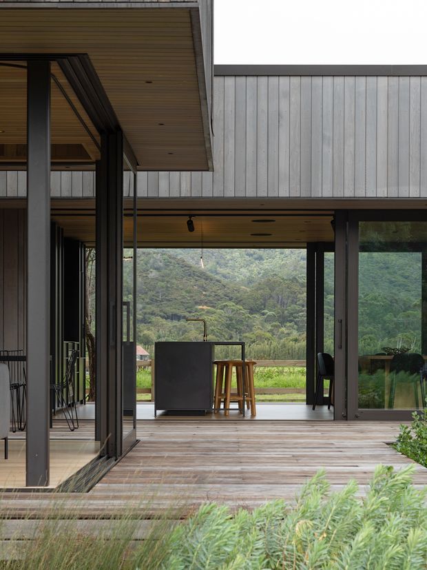 A contemporary pavilion-like home opening onto a timber deck, with extensive dark-framed glazing, vertical pale cladding, and warm timber soffits. A compact outdoor bar or kitchen with stools sits beneath the covered opening, while a lush wooded hillside is visible beyond. The composition emphasizes indoor-outdoor connection and layered architectural volumes.