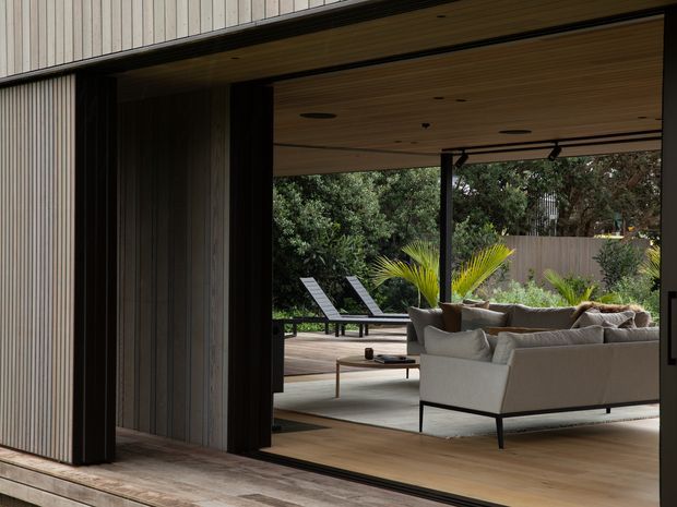 A contemporary indoor-outdoor living area viewed from a timber deck, with wide open sliding glass doors, vertical timber cladding, a warm timber-lined ceiling, and pale timber flooring. The interior contains a large neutral sofa and low coffee table, opening onto a landscaped deck with lounge chairs and planting.
