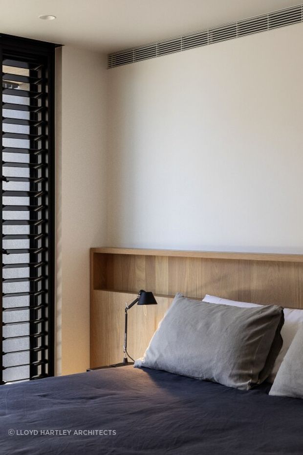 A compact, minimalist bedroom corner with a built-in light timber headboard and integrated shelf, a dark blue-grey bedspread, and neutral bedding. A black adjustable bedside wall lamp is mounted within the headboard, while a tall dark-framed louvred window provides filtered light on the left. A linear ceiling ventilation grille runs across the upper wall, reinforcing the clean contemporary detailing.