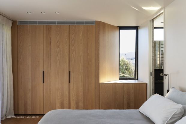 A compact, refined bedroom with a low-profile bed in the foreground and a full-height built-in timber wardrobe along the back wall. Matching timber cabinetry continues beneath a deep window seat beside a tall black-framed window. A partially visible adjoining bathroom or dressing area appears through the open doorway. The room has recessed ceiling lights, pale walls, sheer curtains, and a restrained natural-material palette.