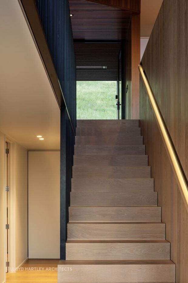 Interior view looking up a modern timber staircase. Broad wood-clad treads rise toward a glazed door and grassy exterior beyond, with a warm wood-paneled wall and integrated linear handrail lighting on the right. A dark vertical slatted screen and matching slatted ceiling frame the stairwell, while a simple hallway with flush doors is visible to the left.