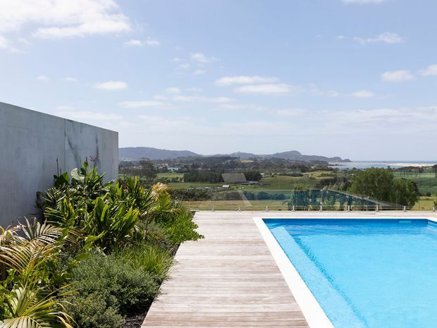 An outdoor pool terrace with a bright blue rectangular swimming pool, pale timber decking, landscaped planting, and a concrete privacy wall. Beyond the glass balustrade is a broad rural landscape with rolling hills and a visible coastal or water horizon under a partly cloudy sky.