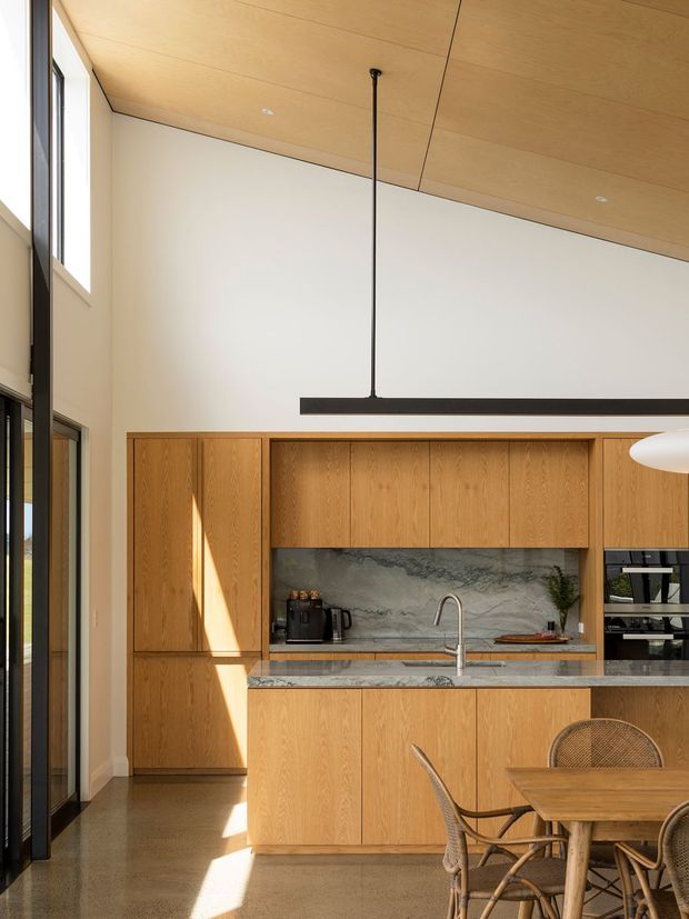 Bright contemporary kitchen and dining area with full-height glazing, warm timber cabinetry, a large island with stone benchtop, integrated appliances, marble-look splashback, and a high timber-lined ceiling. A long black linear pendant hangs over the island, while woven dining chairs and a timber dining table occupy the foreground.