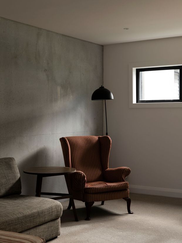 A quiet, moody living room corner featuring a tall brown ribbed upholstered armchair beside a dark round side table and a black arched floor lamp. A grey textured sofa is partially visible in the foreground. The room has a raw grey concrete feature wall, neutral painted walls, carpeted flooring, and a small black-framed horizontal window.