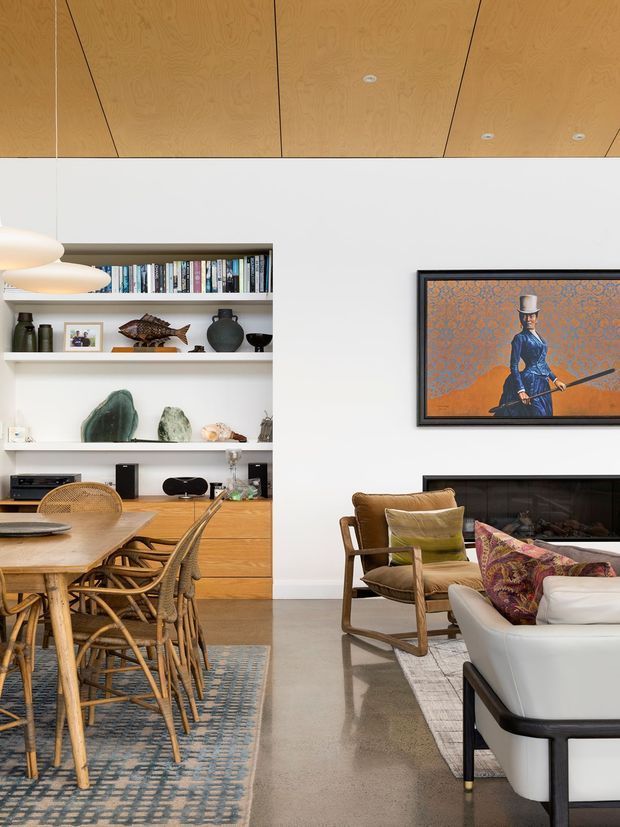 A refined open-plan living and dining interior with a warm timber-lined ceiling, white walls, built-in timber shelving and cabinetry, and polished concrete flooring. The dining area features a timber table with woven rattan chairs, while the living area includes a light upholstered sofa, timber-framed armchair, fireplace, and framed artwork. Decorative shelving displays books, ceramics, sculpture, and other objects.