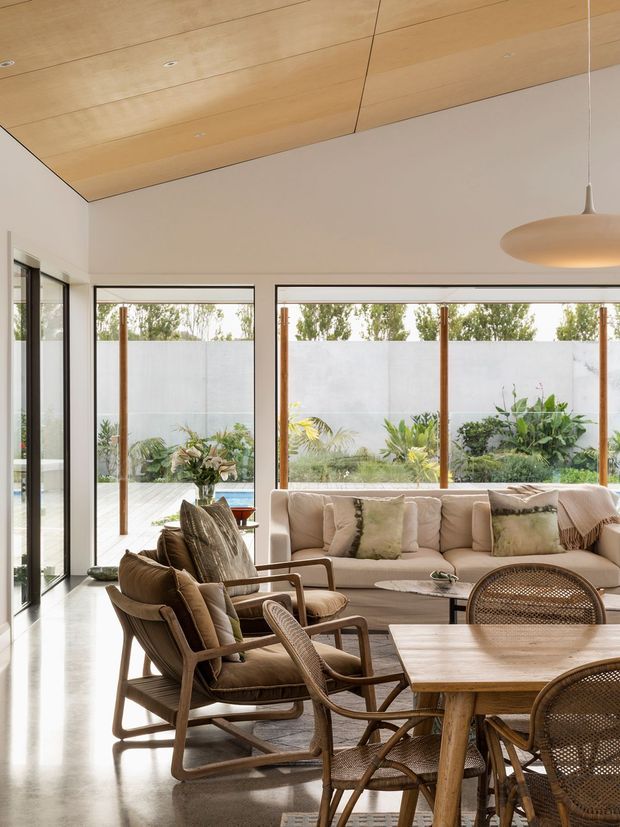 Bright contemporary living room with a warm timber-lined ceiling, polished concrete floor, extensive black-framed glazing and timber mullions overlooking a planted courtyard. The room contains a neutral upholstered sofa, woven lounge chairs, a timber dining table with cane dining chairs, and a large rounded pendant light. The design has a relaxed natural palette and strong indoor-outdoor connection.