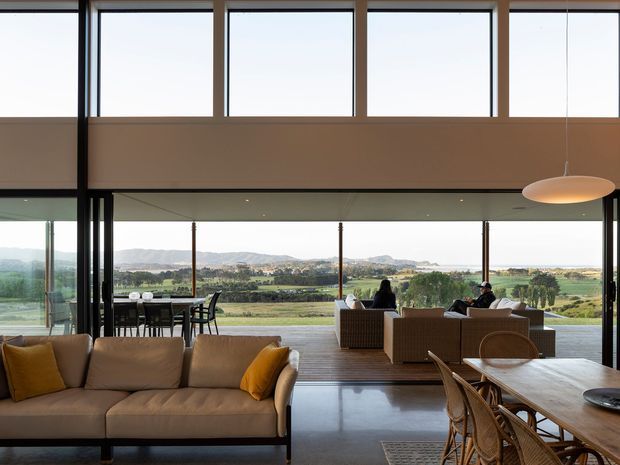 Spacious contemporary open-plan living and dining area with floor-to-ceiling glazing overlooking a rural valley landscape. The room features a large upholstered sectional sofa, timber dining table with woven chairs, a lounge setting near the windows, polished concrete flooring, dark slender window frames, and a suspended oval pendant light.