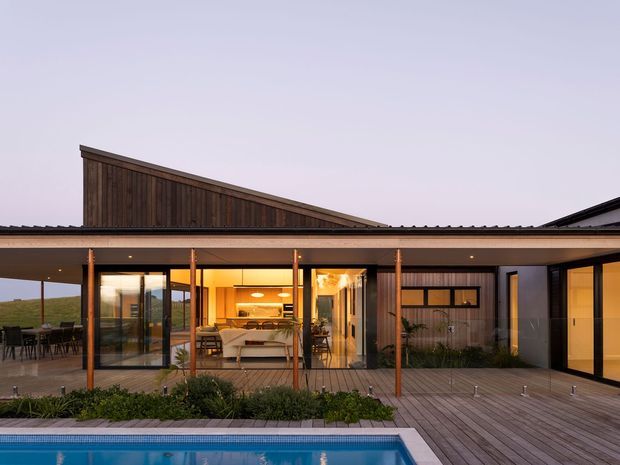 A contemporary single-storey home viewed from the pool deck at dusk. The house has broad floor-to-ceiling glazing, a deep covered outdoor terrace, dark timber cladding, a sloping roof form, and warm interior lighting. Timber decking and a swimming pool occupy the foreground, with an open-plan living and dining area visible through the glass.