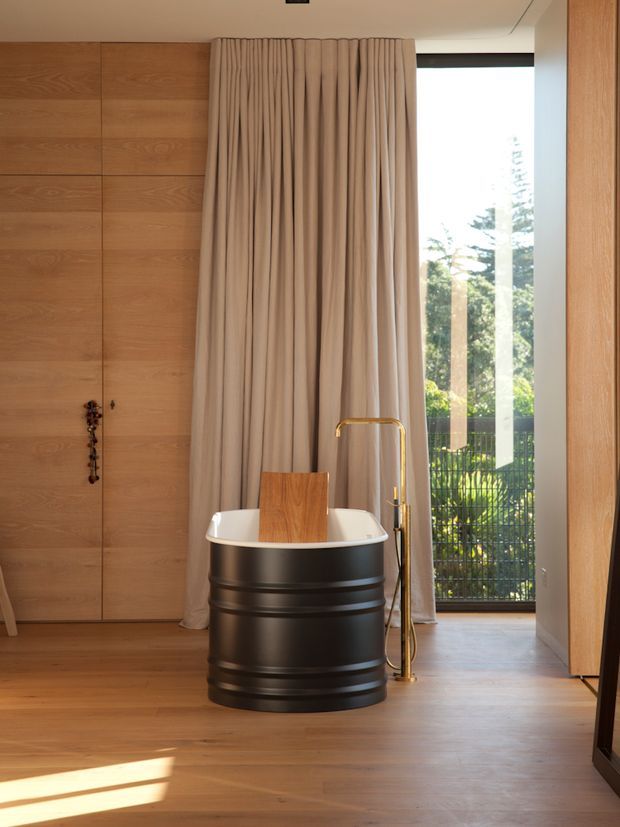 A warm, refined bathroom with a freestanding oval bathtub designed as a dark ribbed barrel-like form, paired with a brass floor-mounted tub filler. The room features extensive warm timber wall panelling and timber flooring, full-height taupe drapery, and a large glazed opening to greenery outside. The composition is minimalist and sculptural, with strong natural light and a calm material palette.