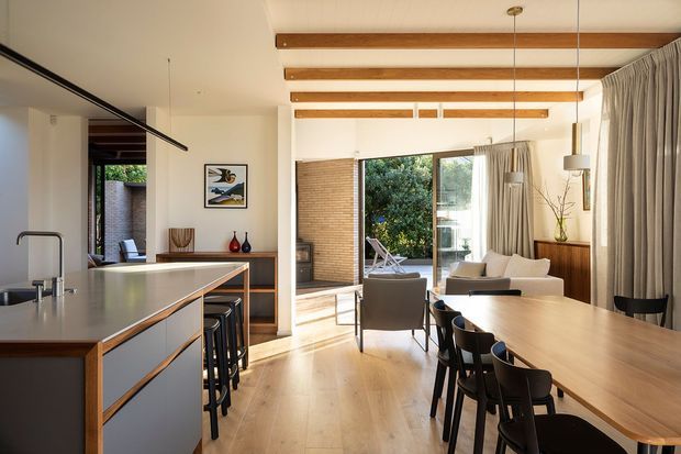A bright, open-plan contemporary living, dining and kitchen space with a long timber dining table, black dining chairs, a minimalist kitchen island with integrated seating, and a lounge area beyond. The room features pale timber flooring, white walls, exposed timber ceiling beams, full-height glazing, wood-and-grey cabinetry, and curtains framing views to greenery.