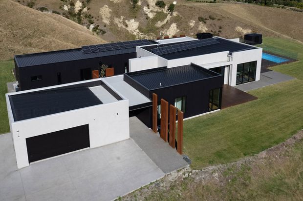 Aerial view of a contemporary, multi-volume single-storey residence on a sloping rural site. The home features crisp white and charcoal-black rendered forms, multiple flat roof sections with dark roofing, rooftop solar panels, large glazed openings, a broad concrete driveway and garage, timber entry screens, and a blue swimming pool visible behind the house.