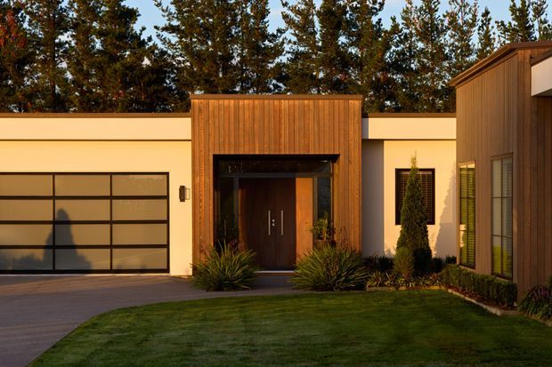 A contemporary single-storey residence viewed from the front, featuring a warm timber-clad entry volume with a dark recessed doorway, pale rendered walls, a wide glazed-panel garage door, and a timber-clad wing with tall windows. Low ornamental planting and a neatly maintained lawn frame the facade, with mature trees behind the property.
