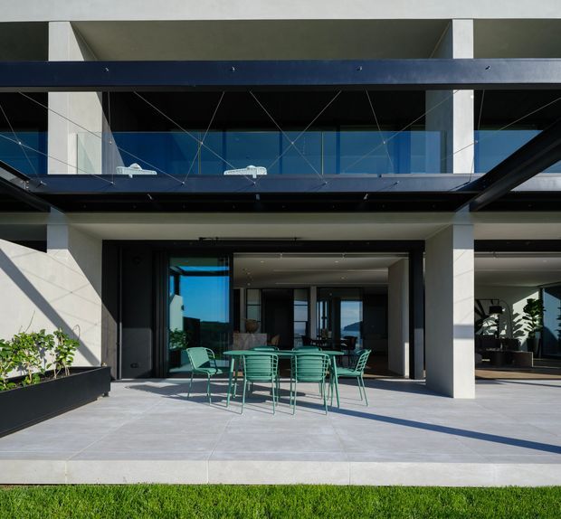Contemporary multi-level building facade viewed from an outdoor terrace, with strong horizontal and vertical structural elements, dark metal framing, extensive glazing, and pale concrete or rendered columns. A paved patio with green outdoor dining furniture sits in front of open glass doors, while indoor furnishings and plants are visible beyond. The image is bright and architectural, emphasizing the relationship between the terrace and the building envelope.