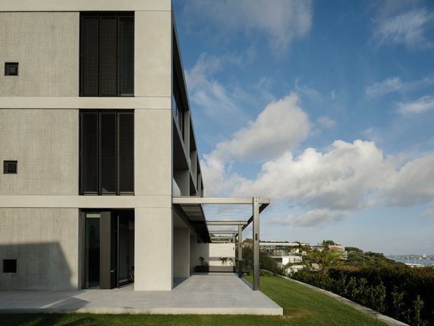 Exterior view of a modern multi-storey residence with a pale textured facade, tall dark louvred window screens, recessed glazing, and a covered outdoor terrace or walkway supported by slim posts. The building sits on a landscaped sloping site with distant suburban and coastal views under a partly cloudy sky.