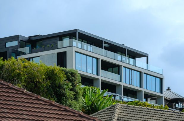 Exterior view of a contemporary multi-storey residential building with a flat roof, strong horizontal and rectilinear massing, extensive floor-to-ceiling glazing, dark cladding, pale rendered or concrete walls, and glass balcony balustrades. Established vegetation and neighbouring tiled roofs occupy the foreground.