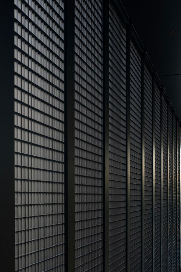 A dark, tightly framed architectural view of a long wall or facade composed of repeated narrow vertical panels and horizontal louvres, divided by substantial dark structural mullions. Dramatic side lighting emphasizes the rhythmic geometric grid and deep perspective, creating an industrial, moody atmosphere.