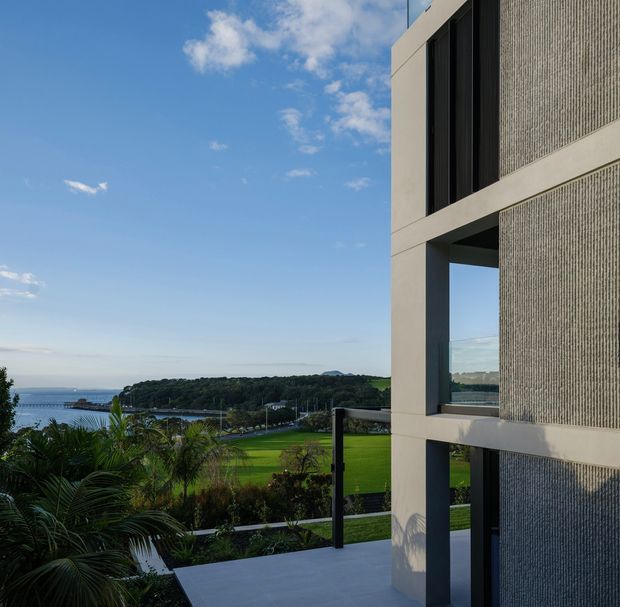 Exterior architectural view of a contemporary multi-storey building with strong rectangular concrete-framed openings, dark vertical glazing, and a finely textured light-grey facade surface. A terrace with glass and dark metal balustrades overlooks landscaped grounds, a waterfront inlet, and a green peninsula under a clear blue sky.
