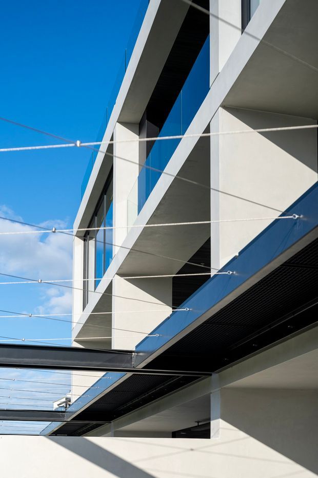 Upward-looking exterior architectural photograph of a modern multi-level building. White rendered or concrete geometric volumes, deep projecting slabs, dark soffits, extensive blue-tinted glazing, and black metal screening create a sharply angular facade. Tensioned horizontal cables span the foreground, with strong sunlight and crisp shadows against a bright blue sky.