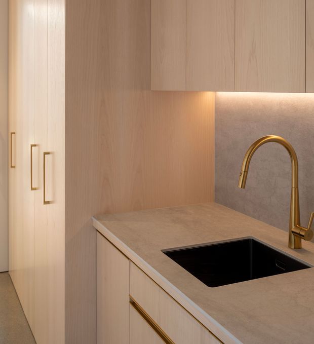 Close-up of a refined contemporary kitchen, focused on a pale timber cabinetry wall, stone-look benchtop, integrated black sink, and curved brass-gold mixer tap. Warm under-cabinet lighting illuminates the grey-beige splashback and work surface, creating a calm, cohesive fit-out.
