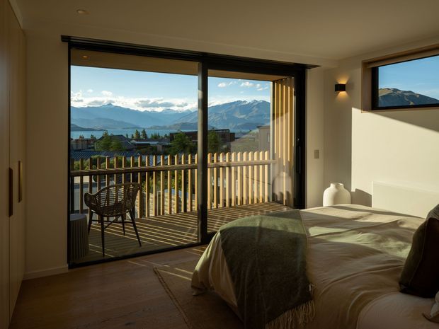 A warm, contemporary bedroom opening onto a timber balcony through large dark-framed sliding glass doors. The room contains a bed with neutral bedding and a green throw, timber flooring, pale walls, a bedside vase, and a small wall light. The balcony overlooks a mountainous landscape and lake or valley, with strong afternoon sunlight entering the room.