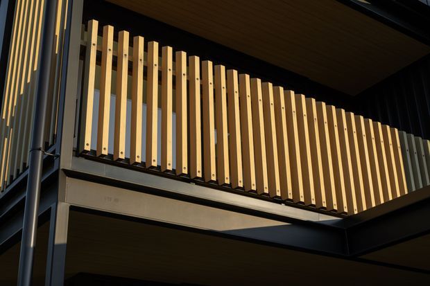 Close-up exterior architectural view of a contemporary building facade, featuring a projecting balcony or screened opening with closely spaced vertical timber battens set against dark metal framing. Dark soffits and structural beams create strong geometric contrast, with warm sunlight highlighting the timber screen.