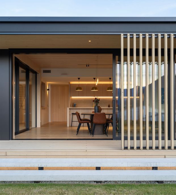 Exterior view into a contemporary open-plan dining and kitchen area through large black-framed sliding glass doors. The facade features a broad dark horizontal roof edge, warm timber soffit and interior finishes, a light timber deck, and a vertical screen of pale timber or metal battens on the right. Inside are a dining table with upholstered chairs, a linear kitchen island, timber cabinetry, pendant lights, and warm integrated shelf lighting.