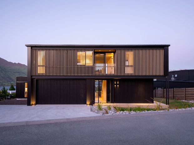 Contemporary two-storey residential facade at dusk, featuring a long rectangular upper level screened by closely spaced vertical timber battens, dark cladding and framing, recessed warm lighting, and large glazed openings. The lower level includes a wide dark garage door, a recessed illuminated entry, and landscaped gravel planting along the concrete driveway. Mountainous terrain and water are visible in the distant background.
