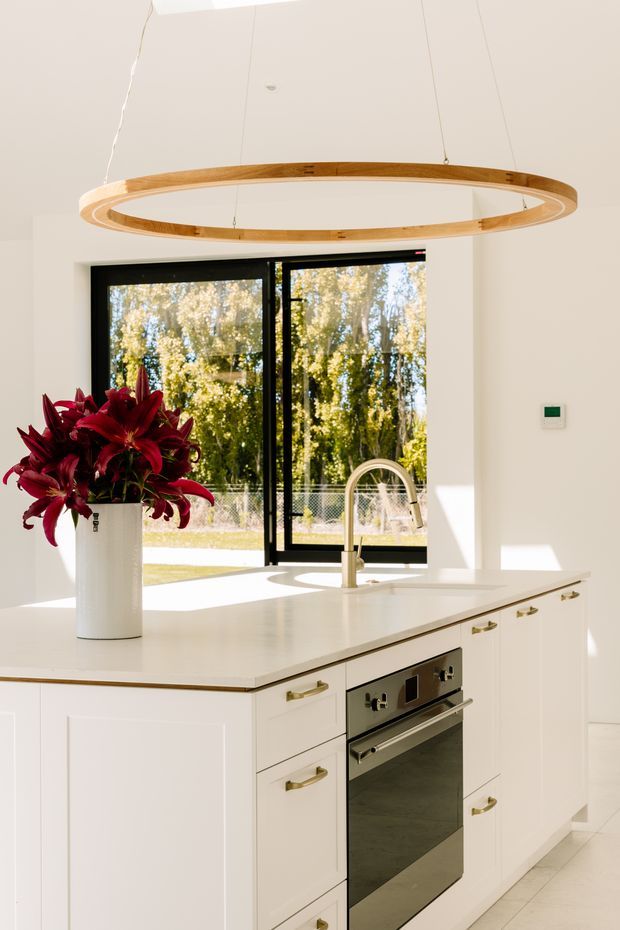 Bright, refined white kitchen centered on a large island with integrated oven, pale benchtop, white shaker-style cabinetry, brass-toned handles, and a curved brushed-metal tap. A large oval timber pendant light hangs above the island. Black-framed glazing opens the room visually to a leafy garden, while a tall white vase of deep red lilies provides a strong accent.