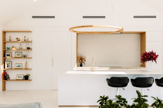 Bright contemporary open-plan kitchen with a large white island and integrated seating, pale timber shelving, concealed white cabinetry, a framed servery or splashback opening, and a sculptural circular timber pendant. Black bar stools and curated shelving provide contrast, while indoor plants and flowers add organic accents.
