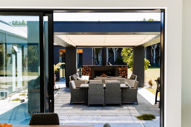 View from inside a contemporary home through large black-framed sliding glass doors to a covered outdoor entertaining area. The patio features a substantial modular outdoor seating arrangement around a central fire feature, a dark feature wall, stone detailing, and a translucent roof canopy. A pool or water feature and additional glazing are visible to the left, with landscaped grounds beyond.