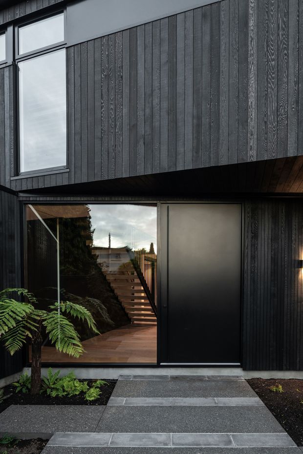 A close exterior view of a contemporary multi-level dwelling with a dark vertically clad facade. Large windows and a glazed entry reveal a timber stair and warm interior beyond. A projecting upper level creates a covered entry zone, while a simple concrete and aggregate path leads through landscaped fern beds to the entrance.
