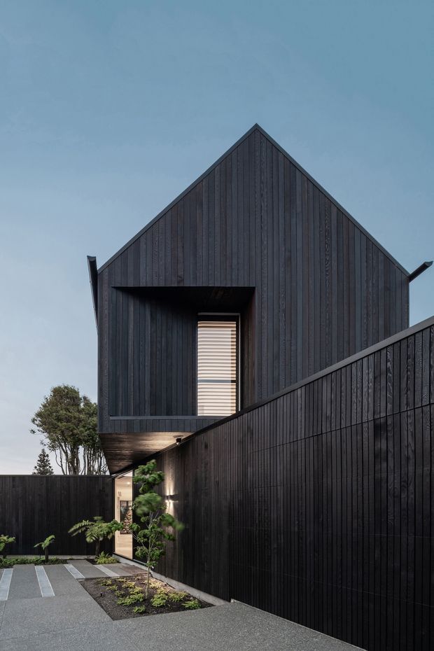 Exterior view of a contemporary gabled house with a dramatic dark vertical timber-clad facade. The building forms a narrow courtyard or entry approach, with a high boundary wall, recessed doorway, small landscaped planting beds, and a paved driveway or walkway. A projecting upper volume contains a deeply recessed opening with a horizontal louvred window screen. The image has a moody, refined architectural character under an overcast sky.