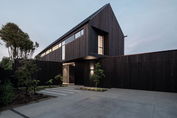 Contemporary dark-clad residential exterior viewed from the driveway, featuring a sharply pitched gable roof, vertical timber cladding, a projecting upper-storey volume, tall horizontal glazing, and a recessed entry. A dark boundary wall and landscaped garden frame the broad concrete approach.