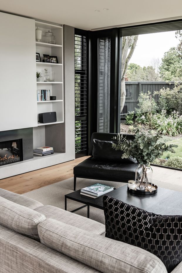 A refined contemporary living room with a pale upholstered sectional, black leather lounge chair, black coffee tables, and a patterned black accent cushion. A built-in white shelving unit and dark fireplace surround line the wall, while full-height black-framed glazing and louvred windows open views to a landscaped garden. Light timber flooring and a neutral palette create a sophisticated, comfortable interior.