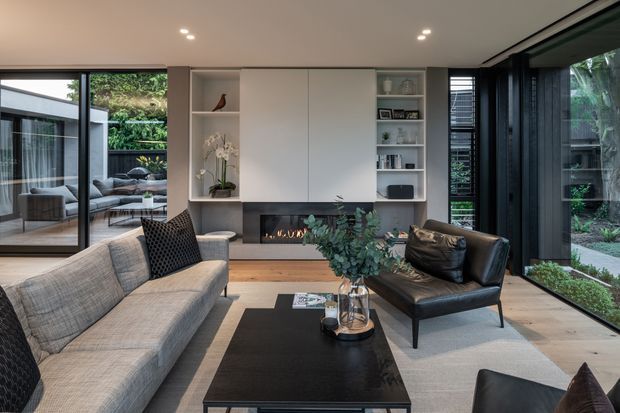Contemporary living room with expansive black-framed glazing, a built-in white media wall and shelving, a linear fireplace, timber flooring, and a refined seating arrangement around a black coffee table. The room opens visually to a covered outdoor terrace and landscaped garden, creating a bright, connected indoor-outdoor atmosphere.