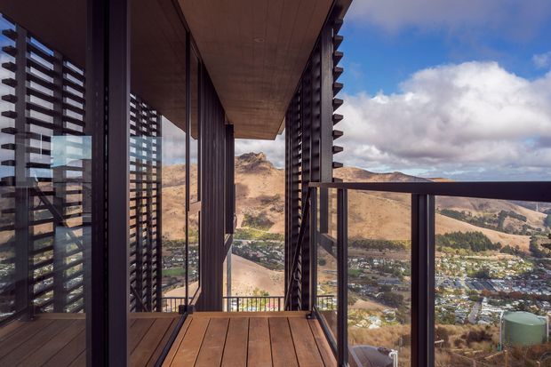 A narrow elevated balcony or terrace walkway beside a modern building, overlooking a dramatic dry mountainous valley and a town below. Dark vertical metal framing and horizontal louvres screen the glazed facade, while the balcony has warm timber decking, a timber soffit, and dark metal balustrades. The image has a contemporary architectural character with strong linear geometry and expansive mountain views.