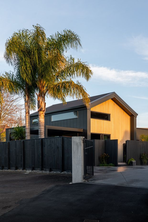 Exterior view of a contemporary two-storey home with a prominent pitched gable roof, dark vertical cladding and fencing, warm timber-toned vertical facade panels, large horizontal windows, and a gated concrete driveway. Palm trees and clear sky frame the residential streetscape.