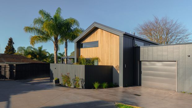 Contemporary single-storey residential house facade with a prominent gable form, vertical timber-clad front wall, dark metal cladding, and an integrated garage. The property is viewed from a paved driveway, with boundary fencing and established palms visible around the home.