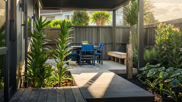 A landscaped outdoor entertaining area viewed from a covered deck, featuring timber decking, a dark slatted overhead roof and fence, a dining table with chairs, built-in bench seating, and dense tropical-style planting in garden beds. The space is warm and sunlit with strong linear shadows and a private courtyard-like character.