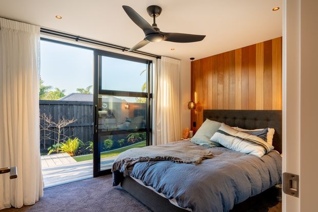 A warm, contemporary bedroom with a low-profile upholstered bed dressed in layered grey bedding, set against a striking vertical timber feature wall. Full-height sliding glass doors with sheer curtains open to a small landscaped courtyard, while a dark ceiling fan and recessed lighting complete the relaxed interior.