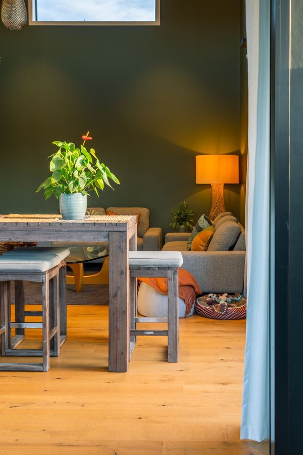 A warmly lit open-plan interior viewed through a doorway, featuring a rustic timber dining table and stools in the foreground, with a grey upholstered sofa, armchair, cushions, orange throw, floor lamp, and indoor plants against a deep olive-green wall. Light timber flooring and abundant natural light create a cosy, relaxed atmosphere.
