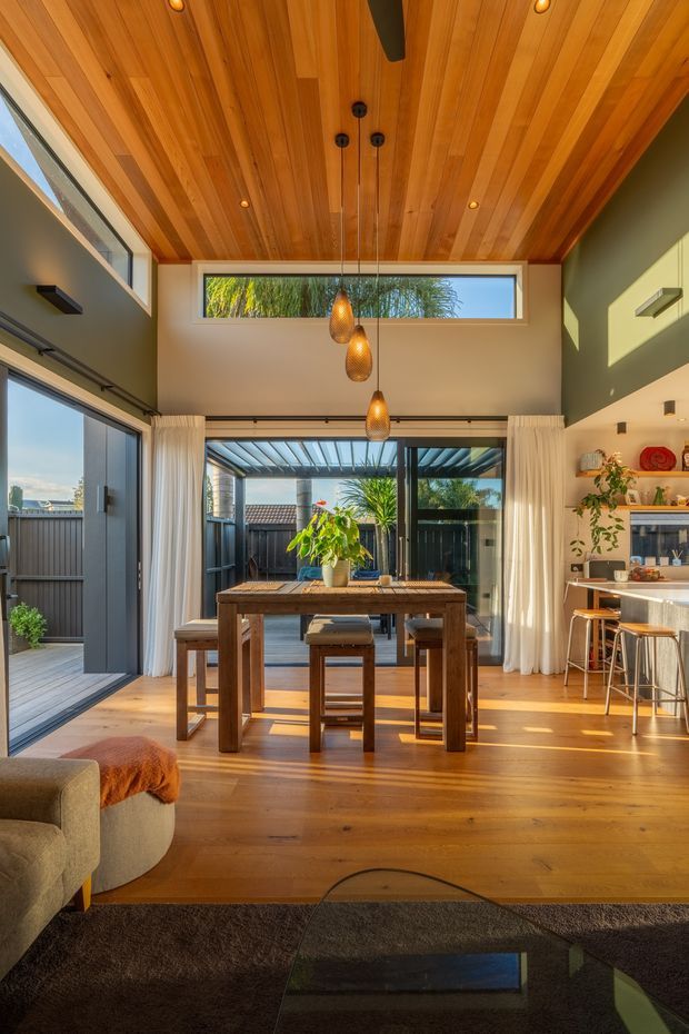 A warm, open-plan living and dining area with a timber-lined vaulted ceiling, polished timber flooring, and large glazed openings connecting to an outdoor patio. A rustic timber dining table and stools sit beneath three woven pendant lights, while a compact kitchen with a peninsula and bar stools is visible on the right. Green-painted walls, sheer curtains, indoor plants, and abundant natural light create a relaxed contemporary interior.