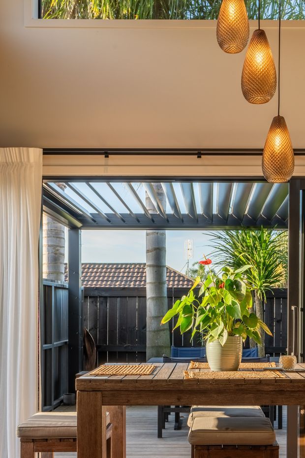 A warm, sunlit dining area opening onto a compact outdoor courtyard or deck. The room features a timber dining table with upholstered benches, sheer curtains, large glazed sliding doors, dark-framed openings, and three woven pendant lights. A pergola with exposed beams extends outside, with tropical plants, a dark timber fence, and a tiled roof visible beyond.