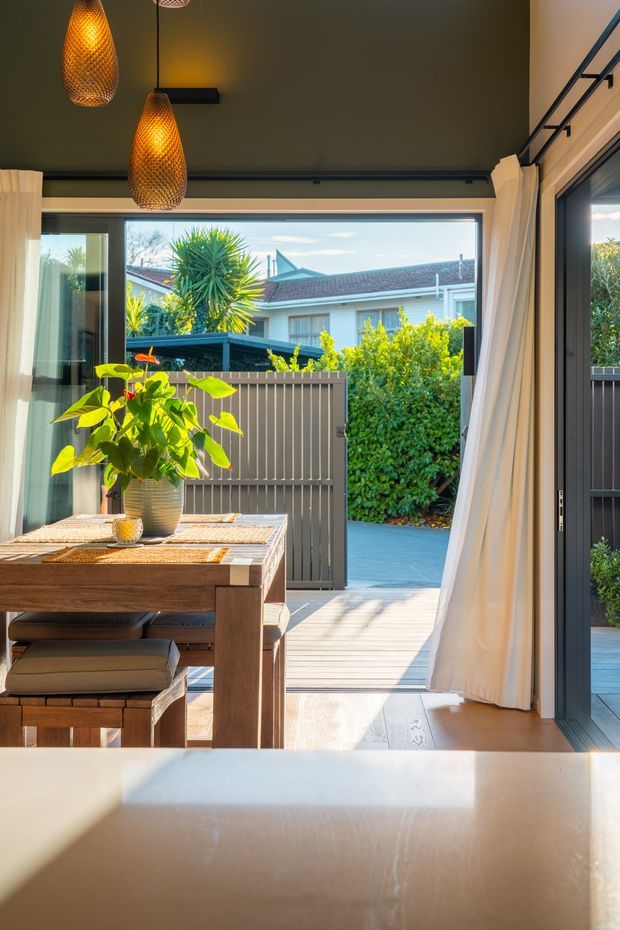 A bright dining area opening directly onto a timber deck and landscaped courtyard through large dark-framed glazed doors. The room features a substantial timber dining table with bench seating, sheer white curtains, a pale stone or composite foreground benchtop, and two woven pendant lights. Warm sunlight contrasts with the dark ceiling and frames, while a neighbouring residential roofline and garden are visible outside.